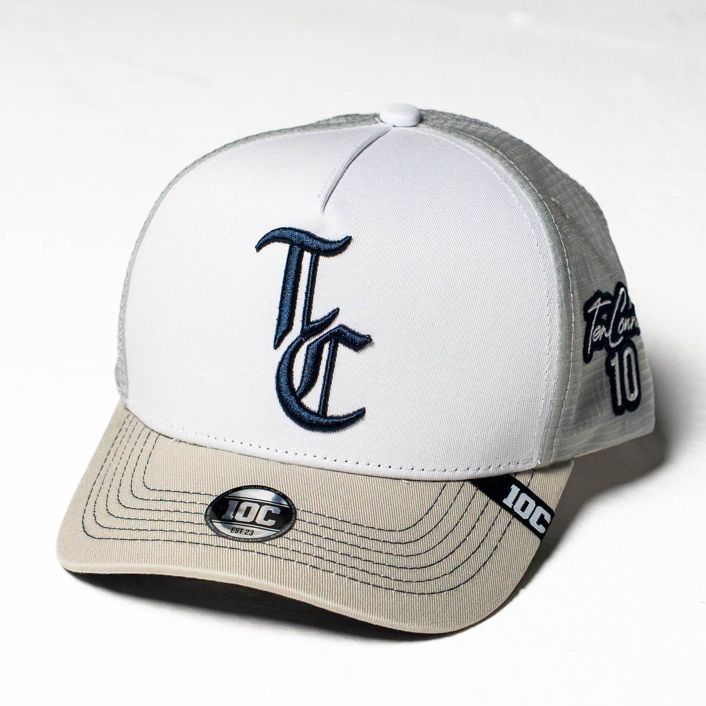 A TC brand trucker cap with a white front panel, grey mesh back, and grey visor. The front features a navy blue embroidered 'TC' logo, and the side displays 'TC' and the number '10'.