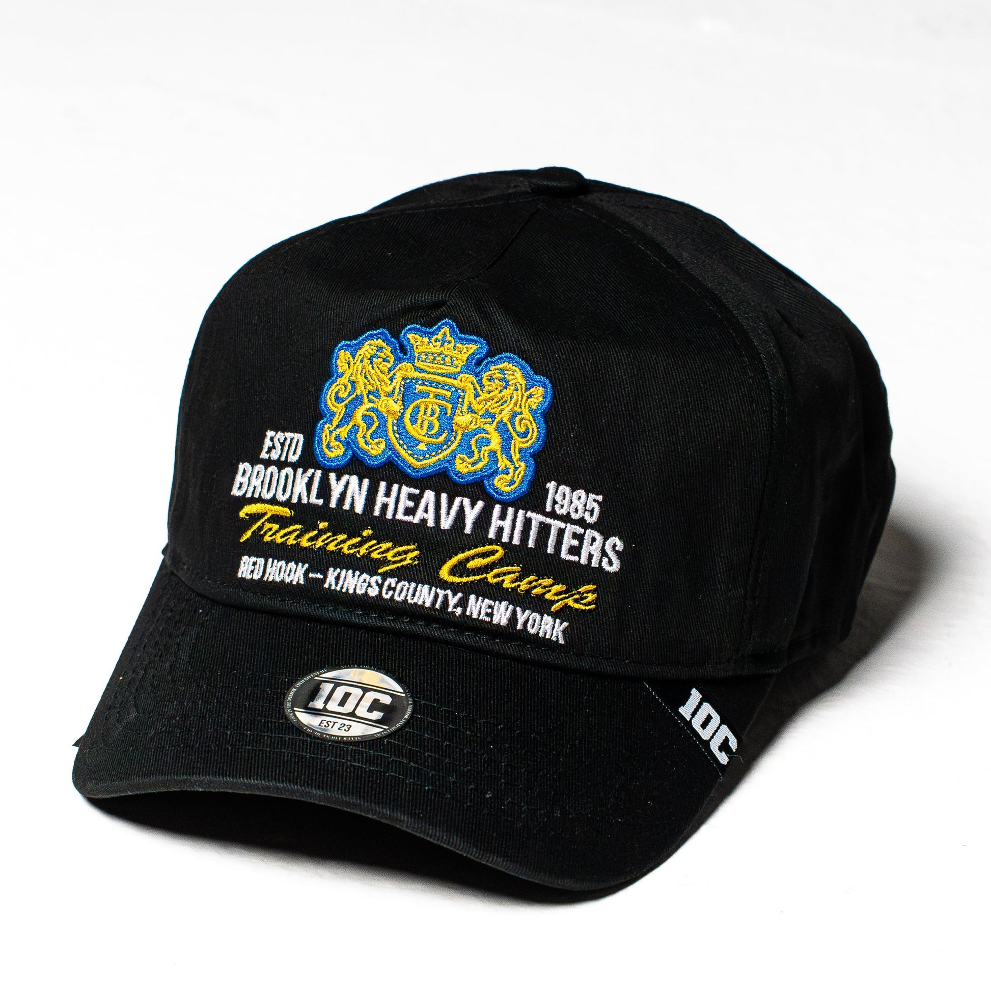 A black relaxed trucker cap featuring embroidered graphics on the front. The design includes a blue and yellow crest with a crown and anchor, alongside white and yellow text reading 'ESTD BROOKLYN HEAVY HITTERS 1985 Training Camp' and 'RED HAWK - KINGS COUNTY, NEW YORK'.