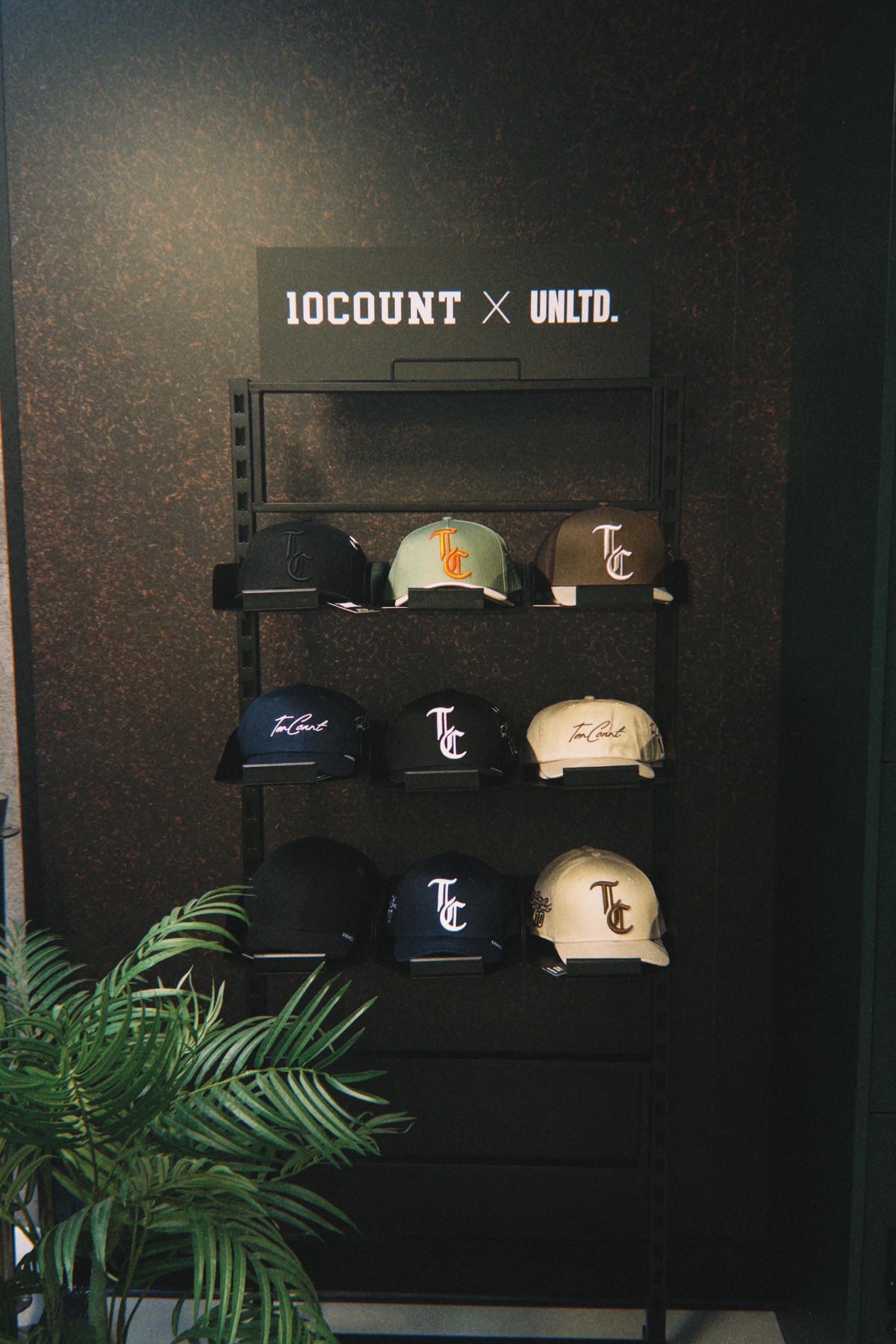 A display rack showcasing multiple 10COUNT x UNLD. collaboration baseball caps in various colors, set against a dark textured wall with a green plant in the foreground.